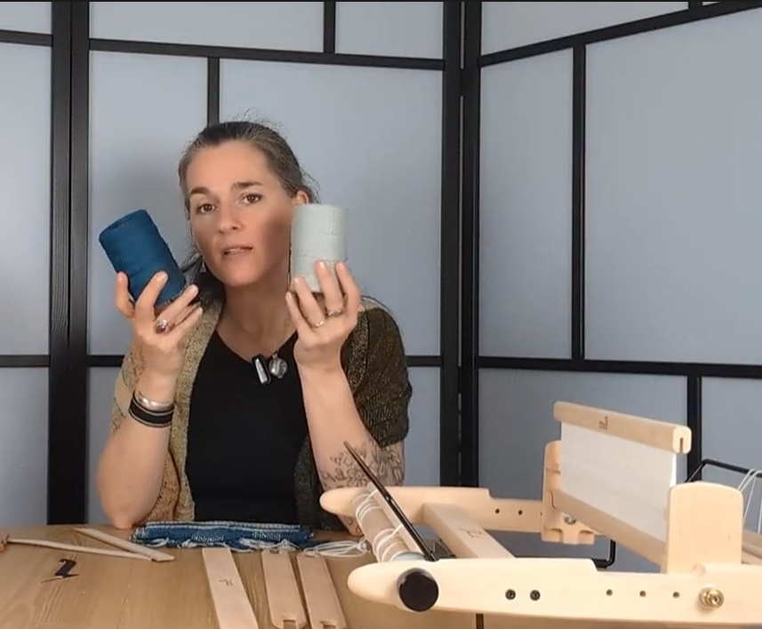 Kursleiterin zeigt zwei Garnrollen im Online-Videokurs Weben auf dem Gatterkamm - mit Webrahmen und Zubehör auf dem Tisch